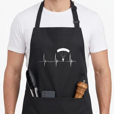Tablier de cuisine noir avec une illustration blanche représentant une ligne de rythme cardiaque intégrant un parachutiste en chute libre.