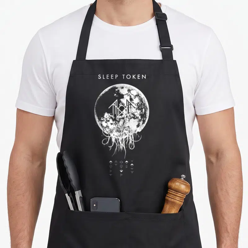 Tablier de cuisine noir avec une illustration blanche représentant une lune et des symboles énigmatiques alignés en bas.