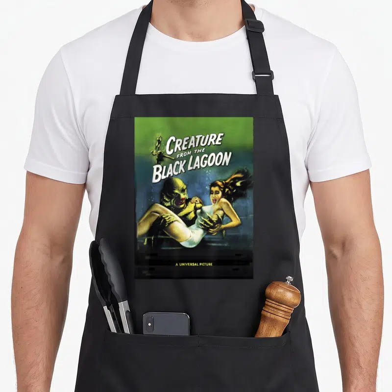 Tablier de cuisine noir avec illustration vintage d'un monstre aquatique tenant une femme sous le titre Creature from the Black Lagoon.