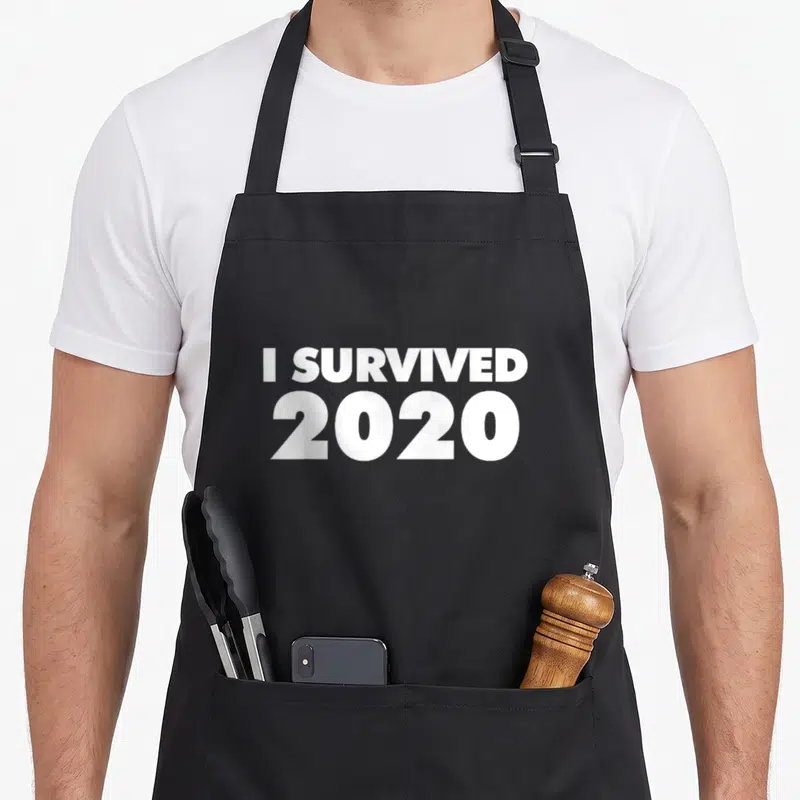 Tablier de cuisine noir avec inscription blanche I Survived 2020 en typographie grasse et minimaliste sur fond uni.