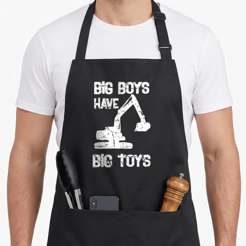 Tablier de cuisine noir avec inscription Big Boys Have Big Toys et silhouette de pelleteuse blanche au centre.
