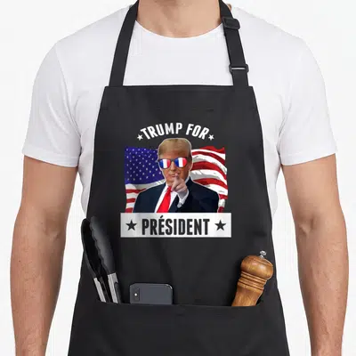 Tablier de cuisine noir avec illustration politique humoristique et drapeau américain en arrière-plan.