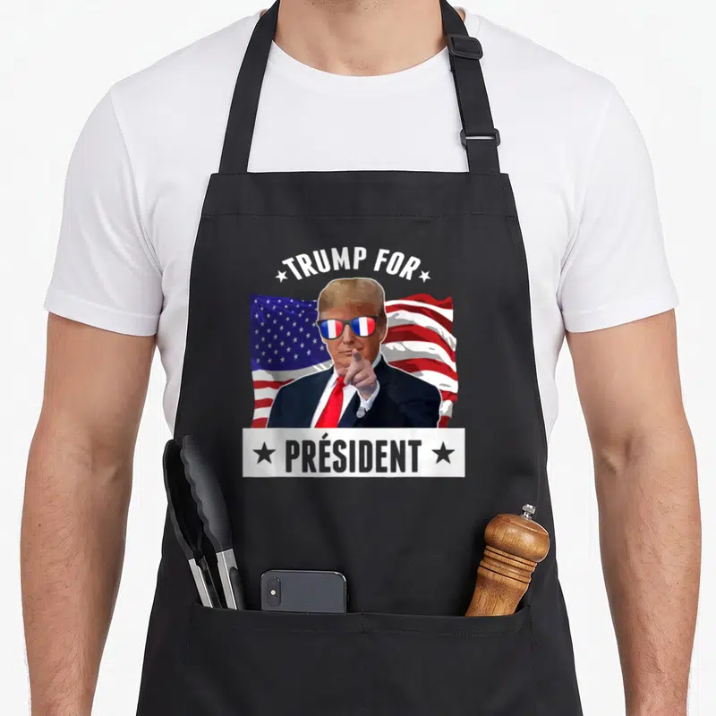 Tablier de cuisine noir avec illustration politique humoristique et drapeau américain en arrière-plan.