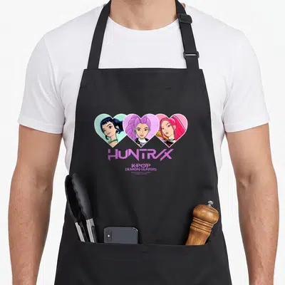 Tablier de cuisine noir avec trois cœurs colorés contenant les visages du groupe Huntrix Hearts et leur nom en violet.