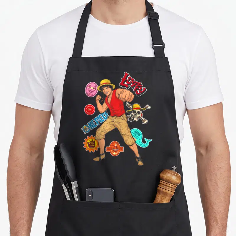 Tablier de cuisine noir avec illustration colorée de Luffy One Piece Stickers et divers logos.