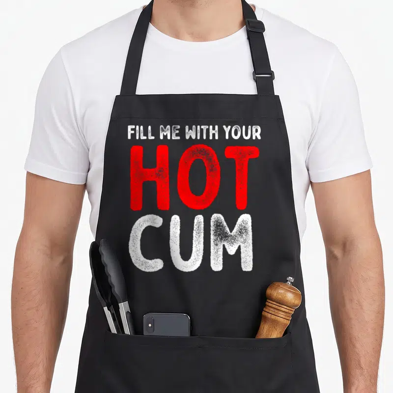 Tablier de cuisine noir avec inscription Fill Me With Your Hot Cum en lettres rouges et blanches texturées.