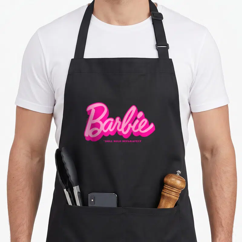 Tablier de cuisine noir avec logo Barbie en rose vif et la mention *Doll Sold Separately en dessous.