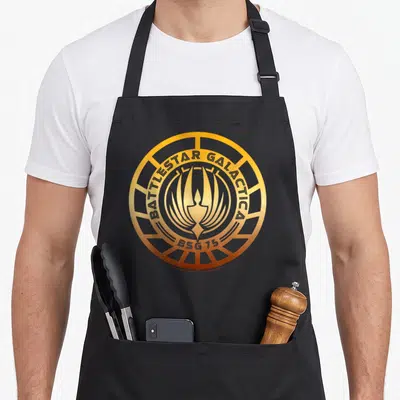 Tablier de cuisine noir avec le logo circulaire doré Battlestar Galactica BSG 75 imprimé au centre.