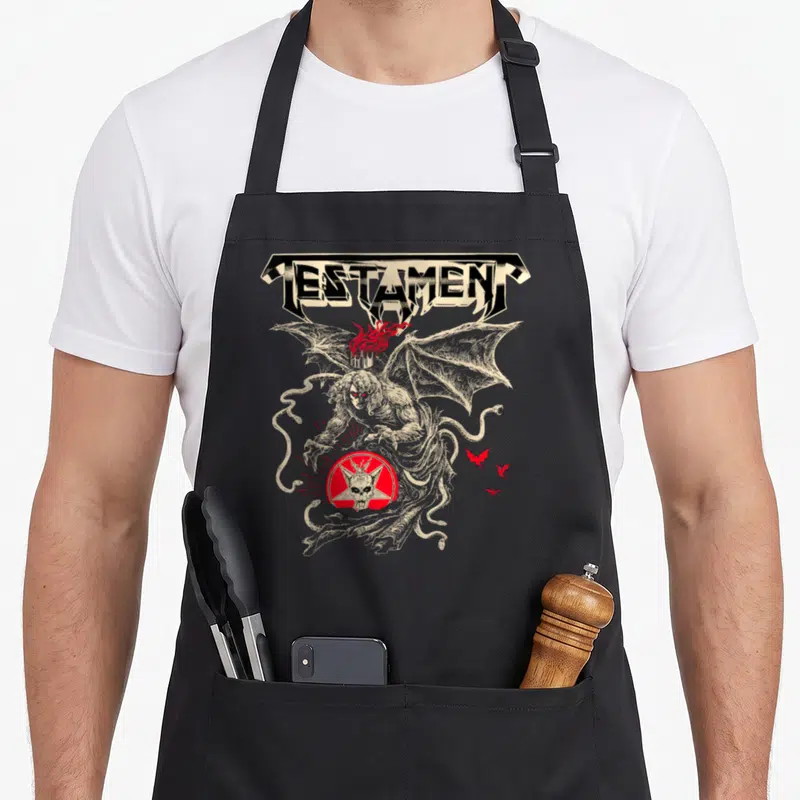 Tablier de cuisine noir avec illustration d'un démon ailé et le logo Testament en typographie métallique.