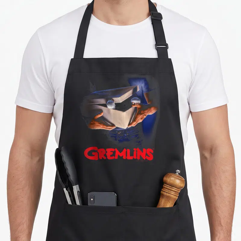 Tablier de cuisine noir avec illustration de mains tenant une boîte contenant deux créatures et le logo GREMLINS en rouge.