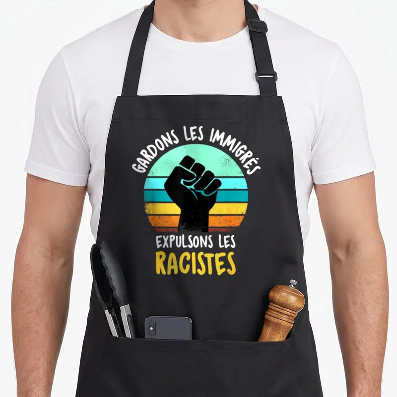 Tablier de cuisine noir avec poing levé et texte Gardons les immigrés expulsons les racistes sur fond circulaire coloré.