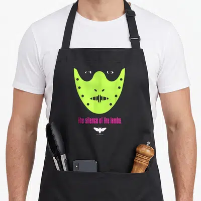 Tablier de cuisine noir avec un masque vert néon et le texte The Silence of the Lambs en rose vif.