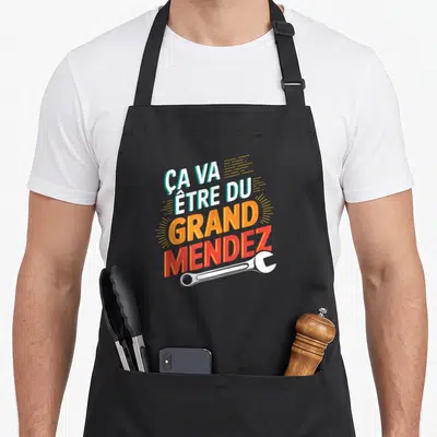 Tablier de cuisine noir avec inscription Ça va être du grand Mendez et une clé anglaise blanche en bas du motif.