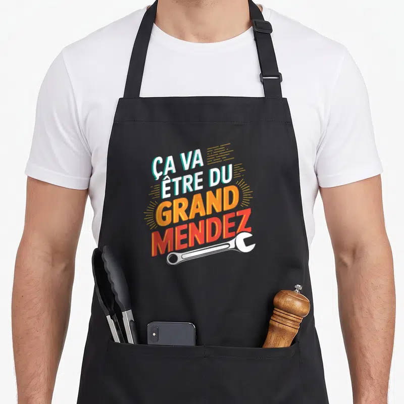 Tablier de cuisine noir avec inscription Ça va être du grand Mendez et une clé anglaise blanche en bas du motif.