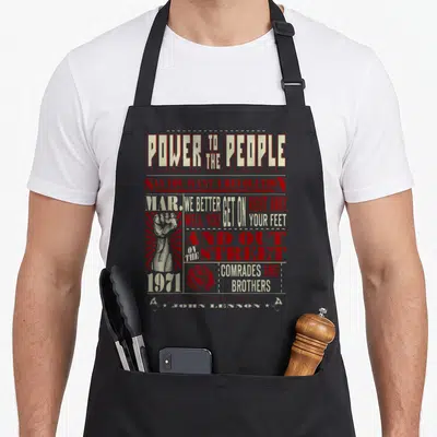 Tablier de cuisine noir avec « Power to the People », poing levé rouge et texte vintage beige sur fond gris uni.