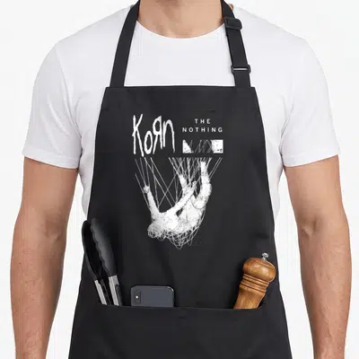 Tablier de cuisine noir avec le logo Korn et une figure blanche suspendue par des fils.