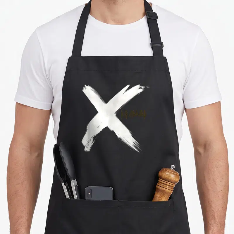 Tablier de cuisine noir avec grand X blanc au pinceau et logo Def Leppard doré sur le devant.