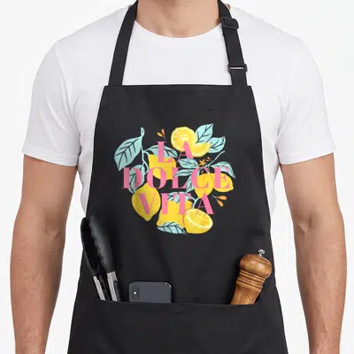 Tablier de cuisine noir avec illustration de citrons jaunes et texte La Dolce Vita en rose pastel sur fond gris.