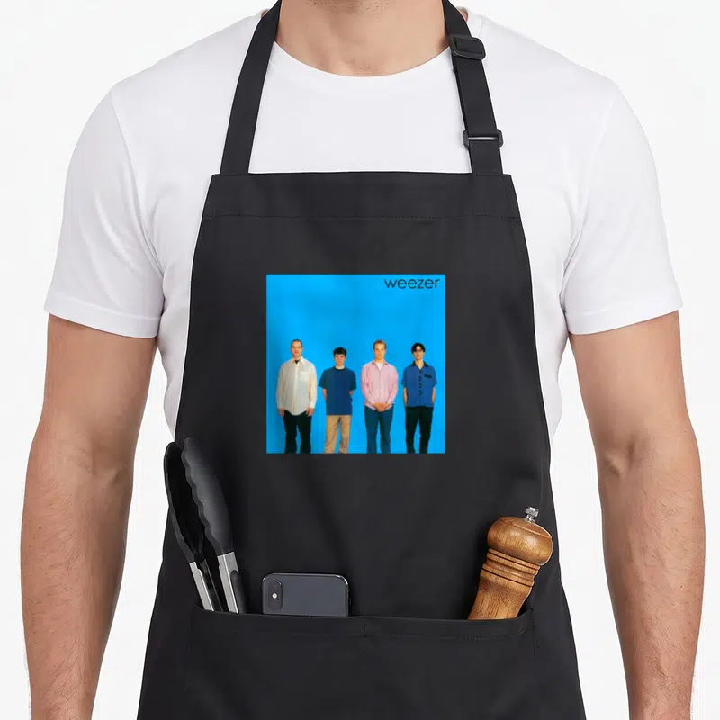Tablier de cuisine noir avec une illustration rectangulaire montrant quatre hommes debout sur un fond bleu uni et le texte weezersur le côté droit.