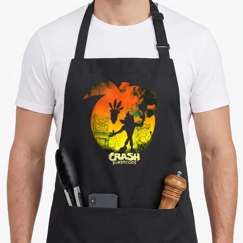 Tablier de cuisine noir avec silhouette de Crash Bandicoot et masques Aku Aku sur fond dégradé orange et jaune.