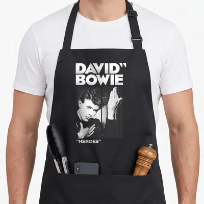 Tablier de cuisine noir avec portrait de David Bowie et texte Heroes en blanc sur fond uni.