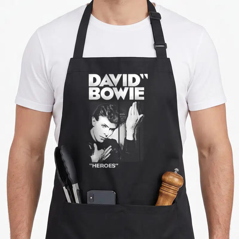 Tablier de cuisine noir avec portrait de David Bowie et texte Heroes en blanc sur fond uni.