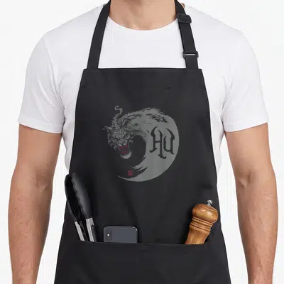 Tablier de cuisine noir avec un dragon aux yeux rouges et le logo The Hu en typographie gothique.