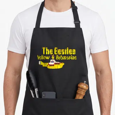 Tablier de cuisine noir avec l'illustration colorée d'un sous-marin jaune et du texte The Beatles Yellow Submarine en lettres ondulées.