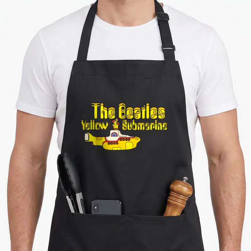 Tablier de cuisine noir avec l'illustration colorée d'un sous-marin jaune et du texte The Beatles Yellow Submarine en lettres ondulées.