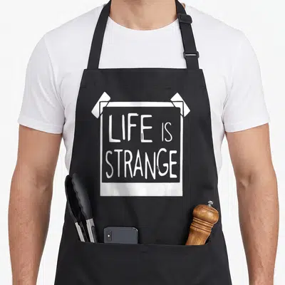 Tablier de cuisine noir avec une illustration blanche représentant un poster collé et le texte LIFE IS STRANGE au centre.