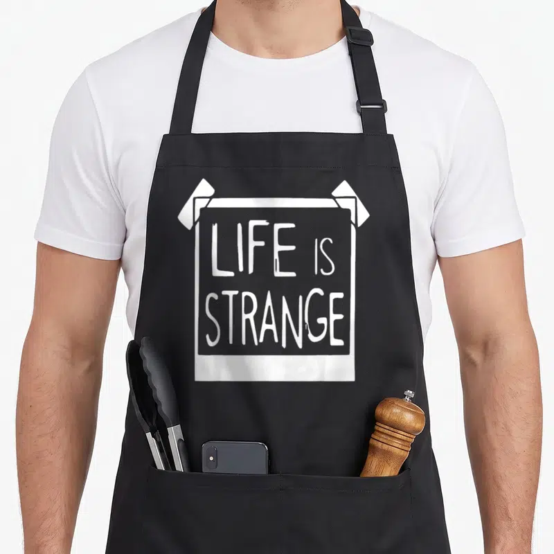 Tablier de cuisine noir avec une illustration blanche représentant un poster collé et le texte LIFE IS STRANGE au centre.