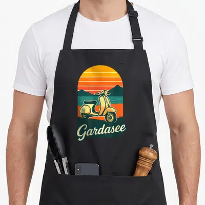 Tablier de cuisine noir avec illustration rétro d'une Vespa jaune et l'inscription Gardasee sous un ciel orange strié.