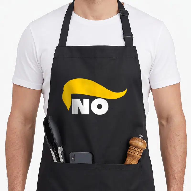 Tablier de cuisine noir avec coiffure blonde stylisée et le mot NO en lettres blanches au centre du motif imprimé.
