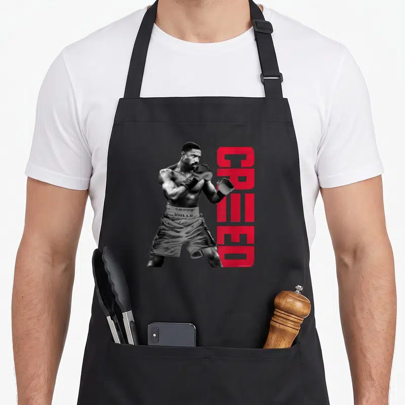 Tablier de cuisine noir avec illustration d'un boxeur en position de garde et texte CREED rouge vertical à droite.
