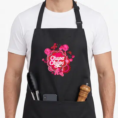 Tablier de cuisine noir orné d'un motif rose vif représentant des bonbons Chupa Chups et des cœurs stylisés.