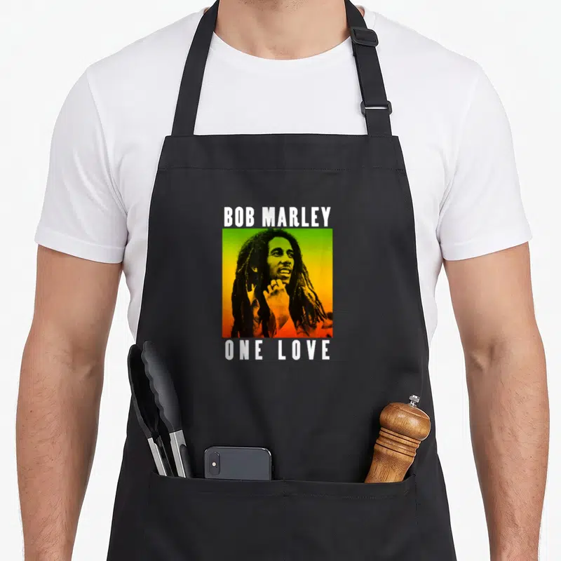 Tablier de cuisine noir avec portrait de Bob Marley sur fond dégradé vert jaune orange et texte blanc Bob Marley One Love.