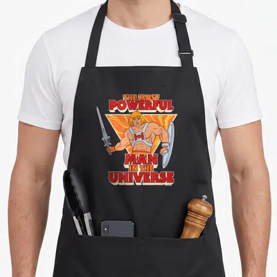 Tablier de cuisine noir avec une illustration colorée de He-Man brandissant un sabre et un bouclier sous un titre en lettres rouges.
