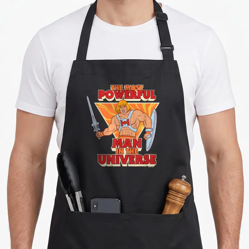 Tablier de cuisine noir avec une illustration colorée de He-Man brandissant un sabre et un bouclier sous un titre en lettres rouges.