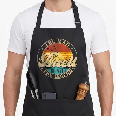 Tablier de cuisine noir avec logo circulaire rétro affichant le nom Buell sur fond de coucher de soleil et d'oiseaux volants.
