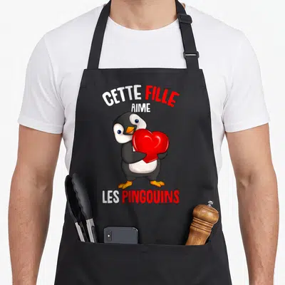 Tablier de cuisine noir avec pingouin tenant un cœur rouge et texte Cette Fille Aime Les Pingouins imprimé en blanc et rouge.