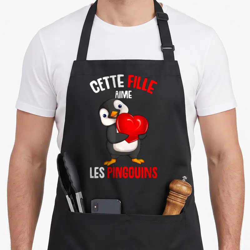 Tablier de cuisine noir avec pingouin tenant un cœur rouge et texte Cette Fille Aime Les Pingouins imprimé en blanc et rouge.