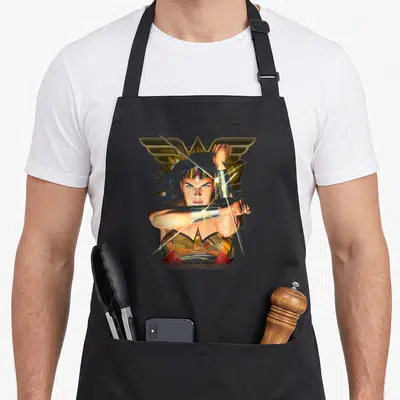 Tablier de cuisine noir avec Wonder Woman croisant les poignets pour défléchir des lances dorées.
