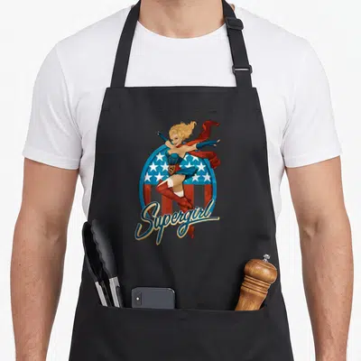 Tablier de cuisine noir avec illustration Supergirl Bombshell blonde en tenue bleue sur fond étoilé américain.