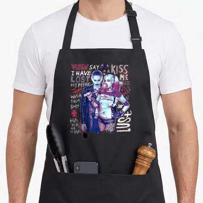Tablier de cuisine noir avec Harley Quinn et Joker entourés de textes colorés et d'un balle de chewing-gum rose.