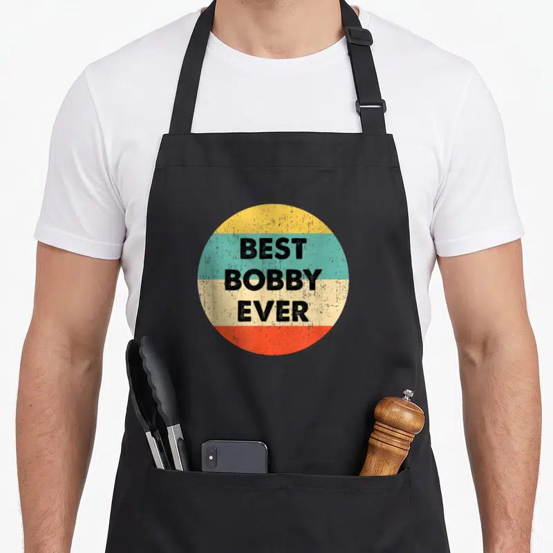 Tablier de cuisine noir avec motif circulaire rétro affichant Best Bobby Ever en lettres noires sur fond multicolore.