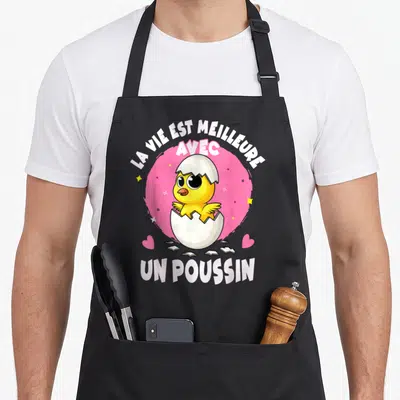 Tablier de cuisine noir avec un poussin jaune sortant d'une coquille sur fond rose et texte blanc.