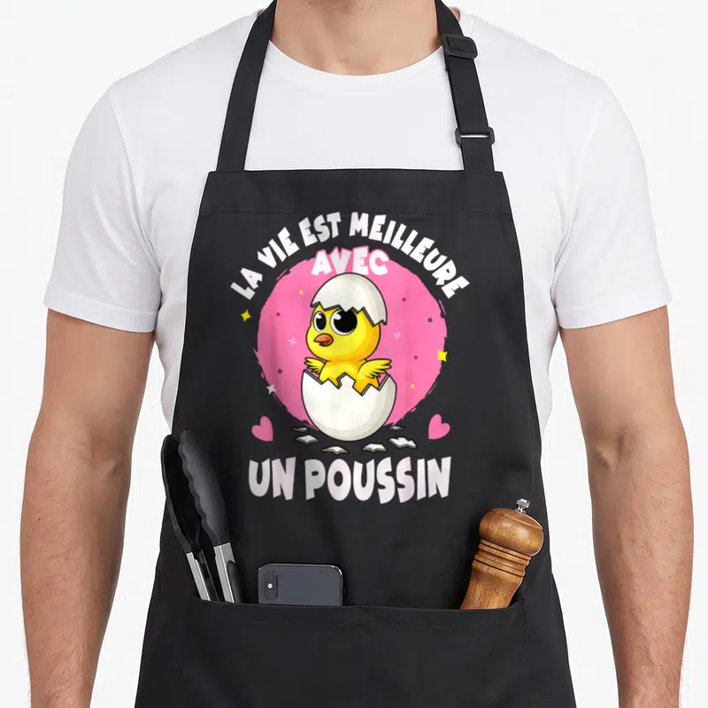 Tablier de cuisine noir avec un poussin jaune sortant d'une coquille sur fond rose et texte blanc.