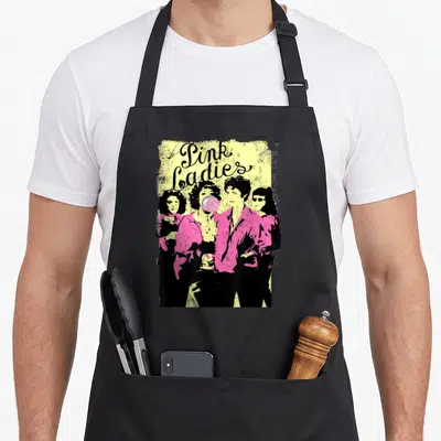 Tablier de cuisine noir avec illustration Pink Ladies en chemises roses et bulle de gomme rose sur fond jaune texturé.
