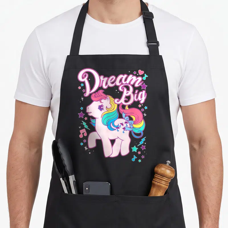 Tablier de cuisine noir avec un poney rose aux cheveux arc-en-ciel et l'inscription Dream Big entourée d'étoiles et de notes de musique.