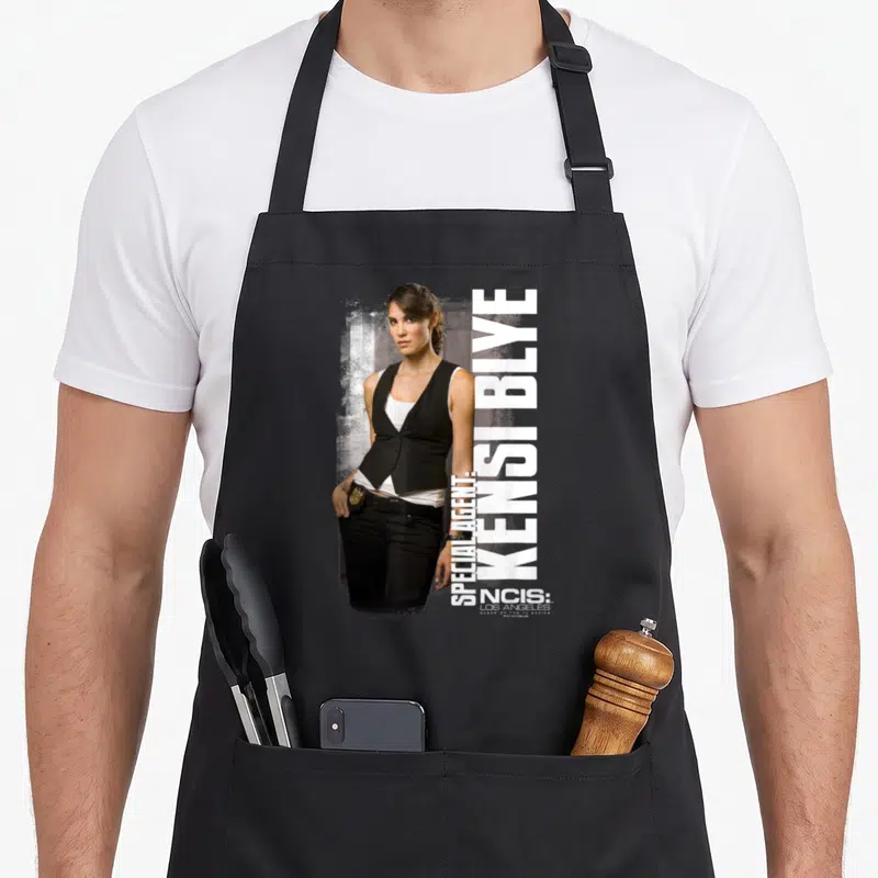 Tablier de cuisine noir avec une illustration de Kensi Blye et le texte Special Agent NCIS Los Angeles imprimé en blanc vertical.